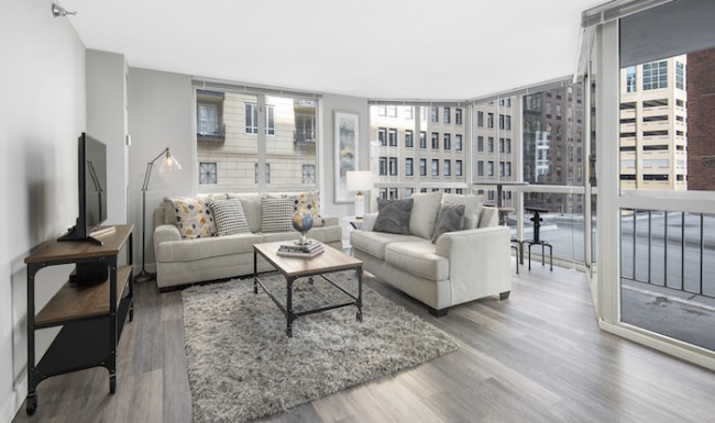 Spacious and well lit living room with wood flooring, large windows and open access to the kitchen.